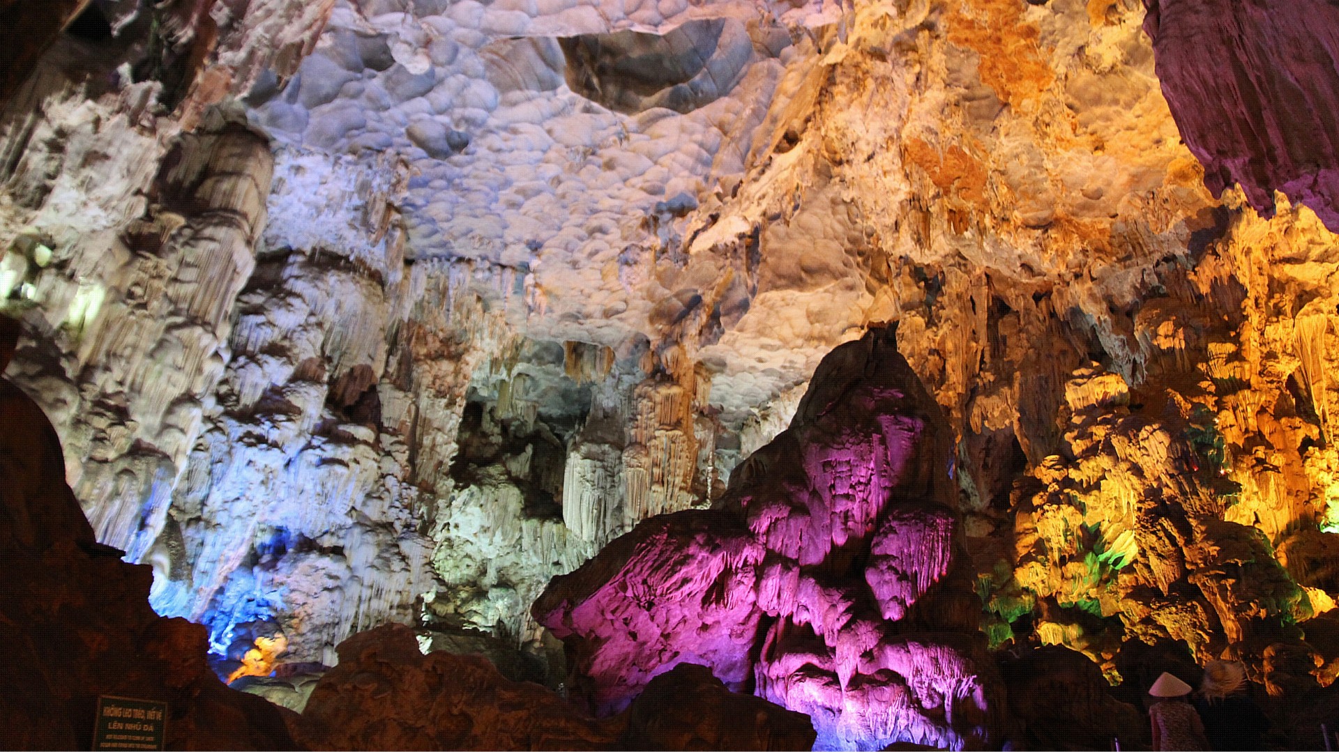 Tourists walk a lit path through a huge limestone cavern with tall stalactite formations overhead.