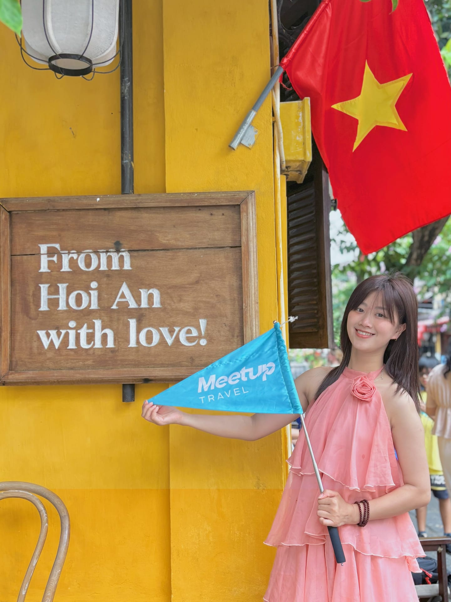 A woman in a pink dress holds a Meetup Travel flag beside a yellow wall with a Hoi An love sign.